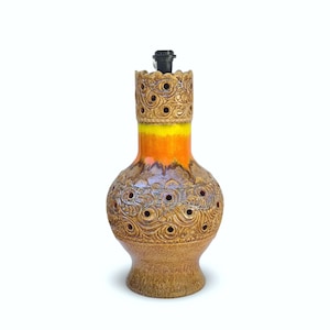 Puede incluir: Una base de lámpara de cerámica con cuerpo bulboso y cuello cilíndrico, decorada con patrones de remolinos en relieve y pequeños recortes circulares. El esmalte transita desde una base marrón texturizada hacia una banda amarilla y naranja.