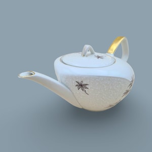 Könnte beinhalten: Weiße Keramik-Teekanne mit goldenem Griff und Ausguss. Die Teekanne hat einen Deckel und ist mit einem gesprenkelten Muster und braunen floralen Designs verziert. Ein klassisches Teezubehör.