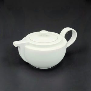 Könnte beinhalten: Eine weiße Keramik-Teekanne mit rundem Korpus, gebogenem Henkel und Ausguss. Der Deckel hat einen kleinen Knopf. Die Teekanne ist zum Aufbrühen von Tee konzipiert und hat eine minimalistische Ästhetik.