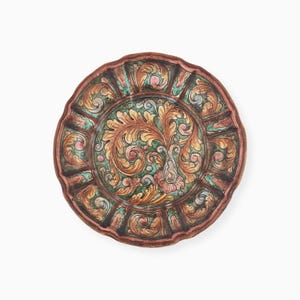 Puede incluir: Un plato de cerámica redondo con borde festoneado, que presenta un medallón central de hojas de acanto de color marrón dorado sobre un fondo verde, rodeado por un borde de motivos florales y de hojas alternos en tonos rosa, verde y marrón.