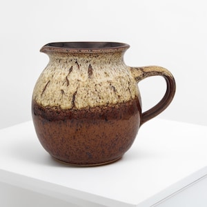 Puede incluir: Una jarra de cerámica con cuerpo redondeado y asa. La jarra tiene una base marrón oscuro, que pasa a un patrón moteado marrón y beige claro hacia la parte superior. El borde y el asa son de color marrón oscuro.