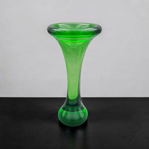Könnte beinhalten: Eine leuchtend grüne Glasvase mit ausgestelltem Oberteil und rundem Boden. Die Vase hat einen schlanken, länglichen Hals und eine glatte, glänzende Oberfläche. Das Design ist schlicht und elegant, geeignet für die Präsentation von Blumen oder als dekoratives Element.