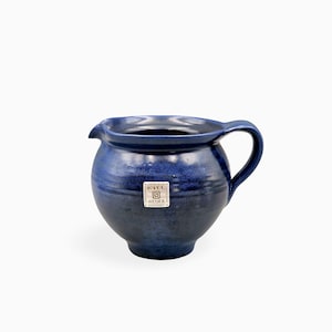 Può includere: Una brocca in ceramica blu profondo con corpo arrotondato, manico curvo e un piccolo beccuccio. Un'etichetta bianca quadrata è applicata sulla parte anteriore, riportante il testo KIEL ATELIER insieme a un grafico a spirale e altro testo.