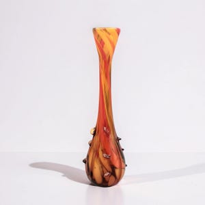 Può includere: Un vaso di vetro alto e sottile con la parte superiore svasata. Il vaso presenta una vivace combinazione di colori arancione, giallo e rosso, con un motivo a spirale. Piccoli accenti di vetro strutturato adornano la parte inferiore. Il vaso è alto circa 30 cm.