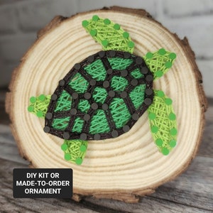 May include: Green and brown string art of a turtle on a wooden slice. The text "DIY KIT OR MADE-TO-ORDER ORNAMENT" is below the turtle.