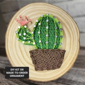 May include: A green string art cactus with a brown pot on a wooden round slice. The text "DIY KIT OR MADE-TO-ORDER ORNAMENT" is on the bottom left corner of the image.