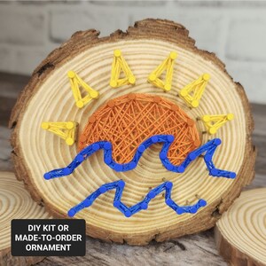 May include: A wooden round ornament with a sun and waves design created with string art. The sun is orange and yellow, and the waves are blue. The text "DIY KIT OR MADE-TO-ORDER ORNAMENT" is at the bottom of the image.