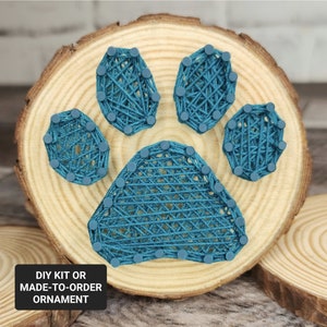 May include: A teal blue paw print string art ornament on a wooden round slice. The ornament is made with nails and string.  DIY kit or made-to-order ornament.