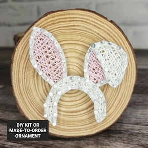 May include: A DIY kit or made-to-order ornament featuring a white and pink string art bunny with silver glitter accents on a wooden round base.  DIY Kit or Made-to-Order Ornament.