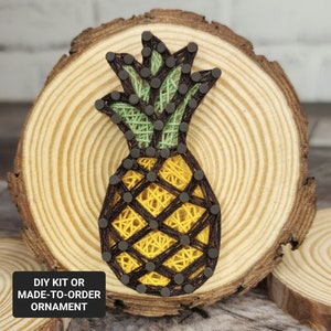 May include: A wooden round slice with a pineapple design created with string art. The pineapple is yellow, brown, and green. The text "DIY KIT OR MADE-TO-ORDER ORNAMENT" is at the bottom of the image.