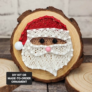 May include: A wooden round ornament with a string art design of Santa Claus's face. The ornament is made with white and red string wrapped around nails hammered into the wood. The ornament has a DIY kit or made-to-order option.