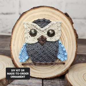 May include: A string art owl ornament on a wooden slice. The owl is made with blue, gray, white, and brown string. The owl has large, black eyes.