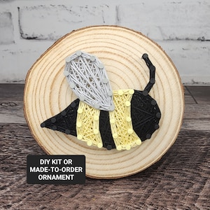 May include: A handmade bee string art ornament on a wooden slice. The bee features black and yellow stripes, a silver wing, and a black stinger. The text "DIY KIT OR MADE-TO-ORDER ORNAMENT" is displayed on a black rectangle.