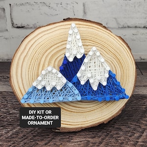 May include: A wooden round slice with three blue and white mountains made with string art. The text "DIY KIT OR MADE-TO-ORDER ORNAMENT" is on the bottom of the slice.