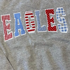 Puede incluir: Una sudadera gris jaspeado de cuello redondo con la palabra EAGLES en letras grandes de tela aplicada. Cada letra muestra un patrón diferente: cuadros vichy rojos y blancos, flores azules, rayas rojas y blancas, motivos botánicos azules, lunares rojos y cuadros vichy azules.