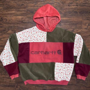 Pode incluir: Um moletom com capuz patchwork com capuz e detalhes em vermelho. O corpo apresenta uma mistura de veludo cotelê verde oliva, estampa floral e painéis bordô. O logotipo da Carhartt está centralizado em um painel vermelho.