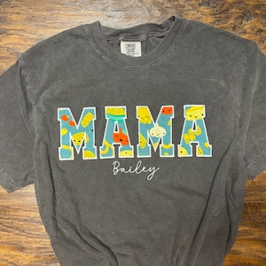 Op de afbeelding: Een donkergrijs T-shirt met het woord "MAMA" in grote witte letters. De letters zijn versierd met een patroon van kleurrijke cartoonvoedingsmiddelen. De naam "Bailey" staat in witte cursieve letters onder het woord "MAMA".
