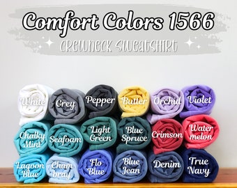 May include: A stack of rolled up crewneck sweatshirts in various colors, including white, grey, pepper, butter, orchid, violet, chalky mint, seafoam, light green, blue spruce, crimson, water melon, lagoon blue, chambray, flo blue, blue jean, denim, and true navy. The text "Comfort Colors 1566 Crewneck Sweatshirt" is at the top of the image.