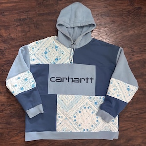 Peut inclure: Un sweat à capuche patchwork bleu clair et bleu foncé avec un panneau central rectangulaire portant le texte carhartt en bleu foncé. Le vêtement intègre des sections matelassées à motif floral et cachemire blanc et bleu sur les manches et le corps.