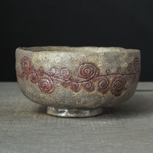 May include: A round ceramic bowl with a textured, mottled grey and brown exterior surface. The bowl features a horizontal band of dark red, hand-painted spiral patterns encircling the middle. It has an uneven, organic rim and a small circular base.