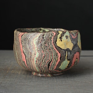 May include: A small, round ceramic bowl featuring a marbled pattern of black, pink, yellow, and light green clay layers. The exterior surface has a textured, uneven finish with a visible rim and a small circular base, showcasing a swirling design.