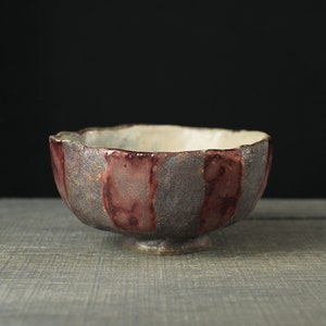 May include: A handmade ceramic bowl with a unique design. The bowl features alternating vertical stripes of dark red and gray-brown. The interior is a light cream color, and the bowl sits on a small, round base.