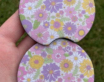 May include: Two round, floral patterned coasters with a scalloped edge. The coasters are made of a dark brown material and feature a white background with pink, purple, and yellow flowers.