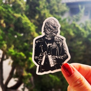 May include: A die-cut sticker featuring a black and white illustration of a creature with scales and fins wearing a leather jacket. The creature is holding a spray paint can in one hand and a bottle in the other, rendered in a high-contrast graphic style.