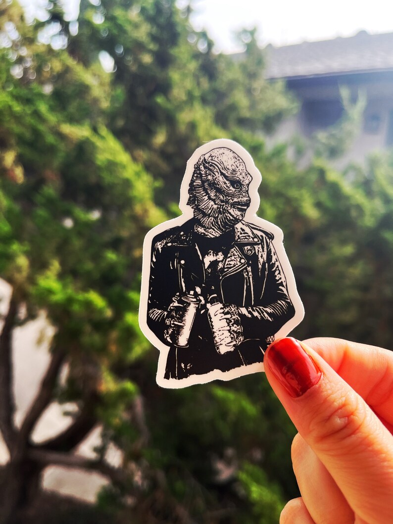 May include: A die-cut sticker featuring a black and white illustration of a creature with scales and fins wearing a leather jacket. The creature is holding a spray paint can in one hand and a bottle in the other, rendered in a high-contrast graphic style.