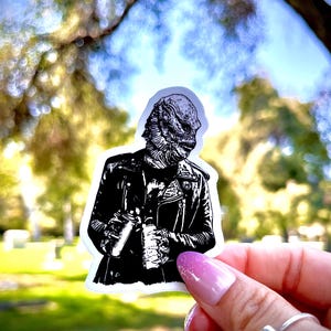 May include: A die-cut sticker featuring a black and white graphic illustration of a creature with textured, scaly skin wearing a leather jacket. The creature is depicted from the chest up, showcasing detailed linework on the face and clothing.