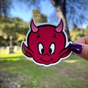 May include: A red devil sticker with a white outline. The cartoon devil has pink horns, ears, and a smiling face with white eyes. The sticker is held up against a blurred green and blue background.