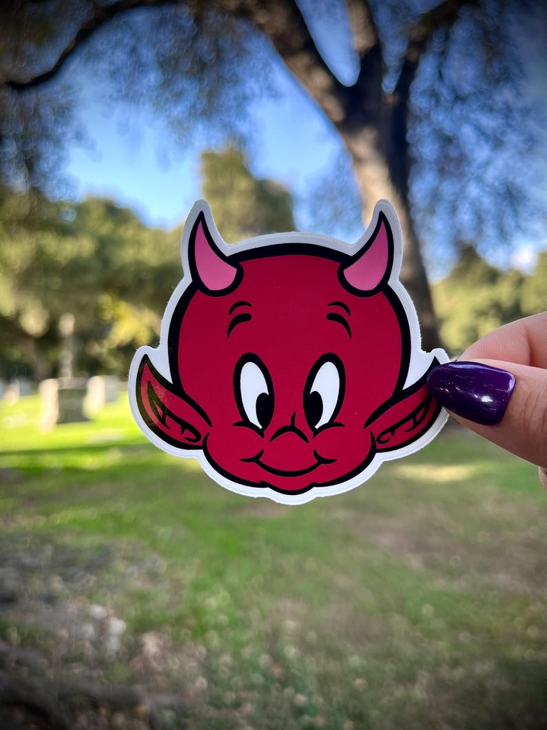 May include: A red devil sticker with a white outline. The cartoon devil has pink horns, ears, and a smiling face with white eyes. The sticker is held up against a blurred green and blue background.