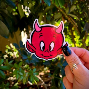 May include: A red devil sticker with pink horns and a smiling face. The sticker is held in front of a green leafy background. The devil sticker is a cartoon style.