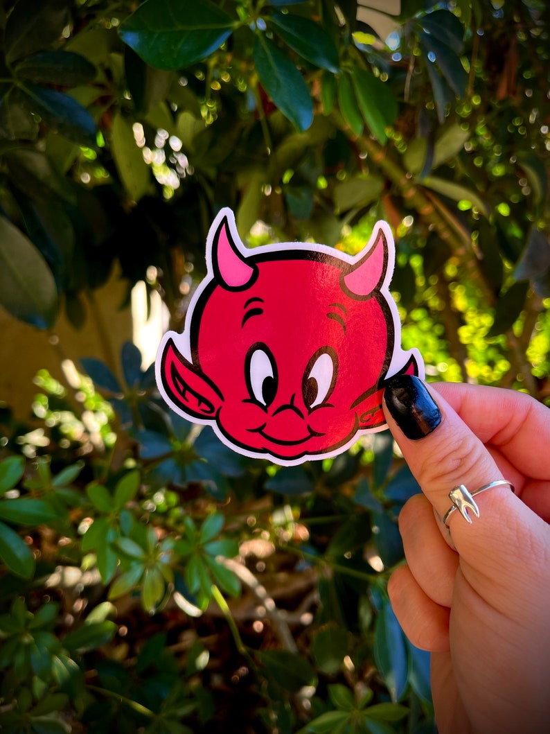 May include: A red devil sticker with pink horns and a smiling face. The sticker is held in front of a green leafy background. The devil sticker is a cartoon style.