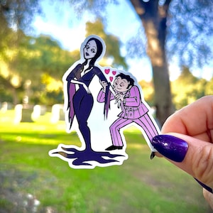May include: A sticker featuring a cartoon illustration of a woman in a black dress and a man in a pink suit. The man is holding the woman's hair, with hearts floating between them. The background is a blurred outdoor scene.