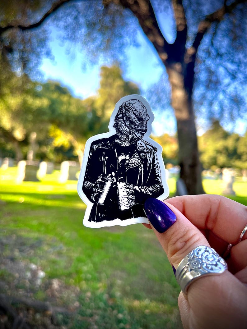 May include: A die-cut vinyl sticker featuring a black and white illustration of the Creature from the Black Lagoon wearing a leather motorcycle jacket and holding two spray paint cans. The design is rendered in high-contrast ink style.