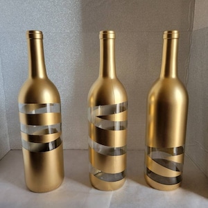 May include: Three gold glass bottles with clear glass sections wrapped with gold tape. The bottles are empty and have a gold metallic finish.
