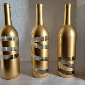 May include: Three gold glass bottles with white stripes. The bottles are empty and have a gold metallic finish.