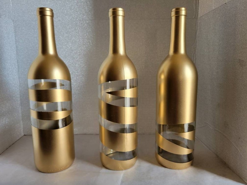 May include: Three gold glass bottles with white stripes. The bottles are empty and have a gold metallic finish.