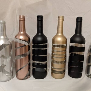 May include: Six glass bottles with different colored paint designs. The bottles are clear glass with a textured surface. The colors are silver, rose gold, black, gold, and black.
