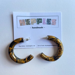 May include: A pair of black and gold hoop earrings with a marbled pattern. The earrings are handmade and have a silver post.