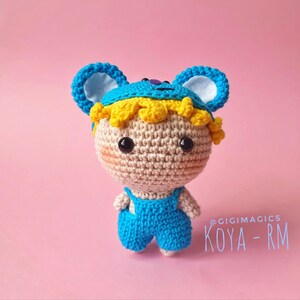 Puede incluir: Muñeca de ganchillo hecha a mano con cabeza de color canela claro, ojos negros y mejillas rosadas. Viste un conjunto azul y un sombrero azul con detalles amarillos. La muñeca está etiquetada "KOYA - RM" sobre un fondo rosa.