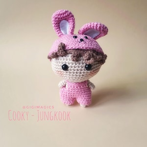 Puede incluir: Muñeca de ganchillo hecha a mano con gorro de conejo rosa y mono. La muñeca tiene una cara beige, ojos negros y pelo castaño. El texto "COOKY - JUNGKOOK" está escrito en rosa en la parte inferior. Un artículo hecho a mano.