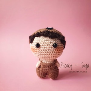 Puede incluir: Muñeca de ganchillo con cabeza redonda, ojos negros y mejillas rosadas. La muñeca lleva gorra y peto marrones. El fondo es de color rosa liso. El texto de la derecha dice "Shooky - Suga @GIGIMAGICS".