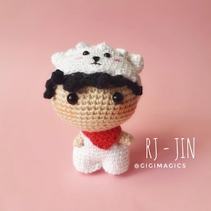Puede incluir: Muñeca de ganchillo hecha a mano con cabeza beige, pelo negro y un sombrero blanco con cara de oso. La muñeca lleva una bufanda roja y un mono blanco. El texto "RJ - JIN" y "@GIGIMAGICS" son visibles sobre fondo rosa.
