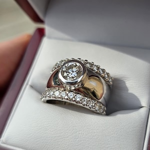May include: A diamond ring with a large, round center stone and smaller diamonds set in a band. The ring is in a maroon jewelry box with a white interior. The ring is made of silver and gold.