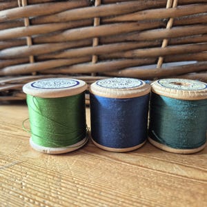May include: Three wooden spools of thread in shades of olive green, navy blue, and dark teal. Each spool features a circular paper label on the top with printed text. The spools are arranged in a horizontal row, showcasing the vibrant thread colours.