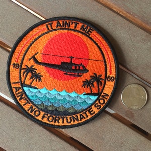 May include: Embroidered patch with a sunset design featuring a helicopter, palm trees, and ocean waves. The patch has the text "IT AIN'T ME" and "AIN'T NO FORTUNATE SON" in a circular pattern, with the numbers "19" and "69".