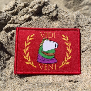 Puede incluir: Una taza de cerámica blanca con asa negra y el texto "But First, Coffee" en negrita negra. La taza se encuentra sobre un fondo borroso, sugiriendo un ambiente acogedor.