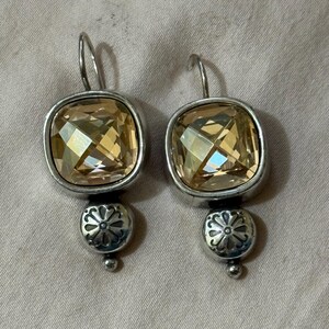 May include: A pair of drop earrings featuring square-cut, light amber-colored faceted crystals set in silver-toned metal. Below each crystal is a small, round silver-toned bead with an embossed floral pattern and a tiny spherical tip at the base.